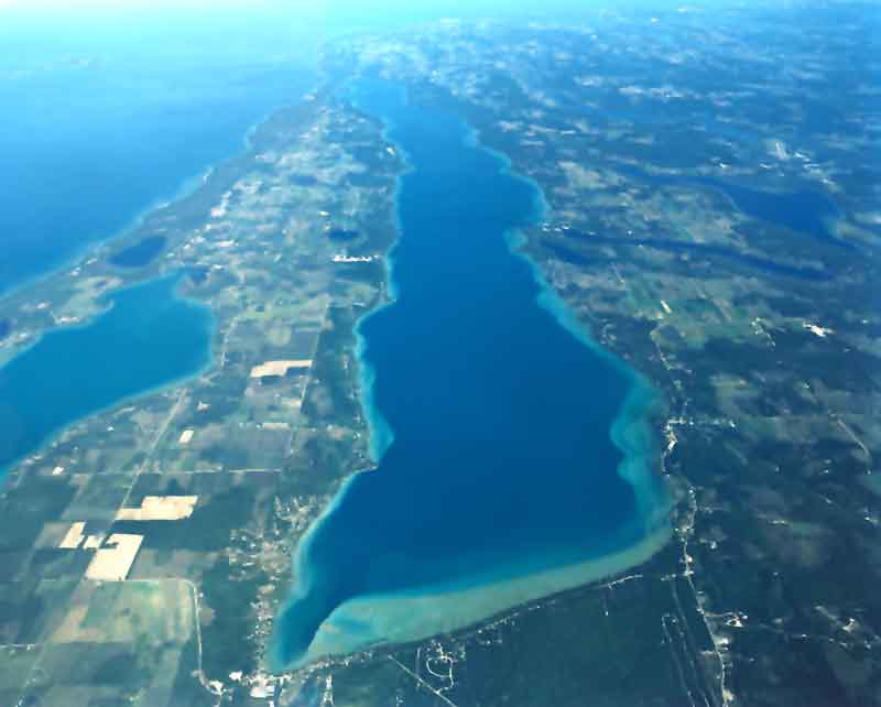 Aerial view of Torch Lake looking north, with deep blue water in the center and turquoise shallows along the shore and across the south end